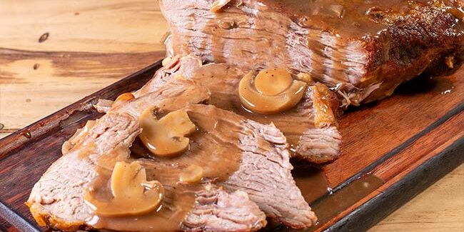 Carne Assada ao Molho com Champignon para Viagem Campinas – Ramalho Carnes Carne assada fatiada e suculenta coberta com molho madeira e champignon, servida em tábua de madeira. Opção de carne assada para viagem em Campinas da Ramalho Carnes.