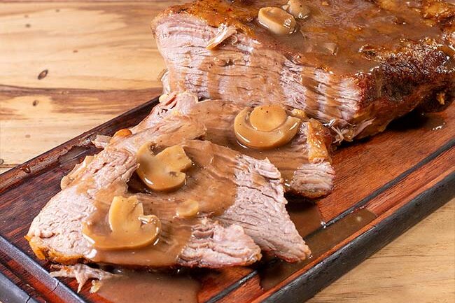 Carne assada fatiada e suculenta coberta com molho madeira e champignon, servida em tábua de madeira. Opção de carne assada para viagem em Campinas da Ramalho Carnes.