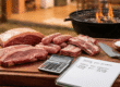 Tábua de madeira com peças cruas de picanha, linguiça e costela, ao lado de uma calculadora e um caderno com anotações sobre como calcular carne para churrasco.
