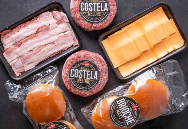 Kit Hambúrguer Artesanal Costela e Bacon Vista superior dos ingredientes do kit hambúrguer artesanal contendo hambúrgueres de costela nelore, fatias de bacon, queijo e pães brioche.