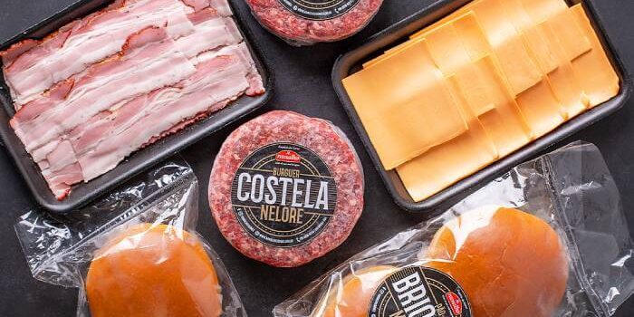 Vista superior dos ingredientes do kit hambúrguer artesanal contendo hambúrgueres de costela nelore, fatias de bacon, queijo e pães brioche.