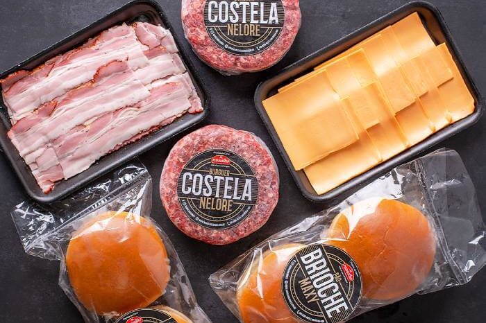 Vista superior dos ingredientes do kit hambúrguer artesanal contendo hambúrgueres de costela nelore, fatias de bacon, queijo e pães brioche.