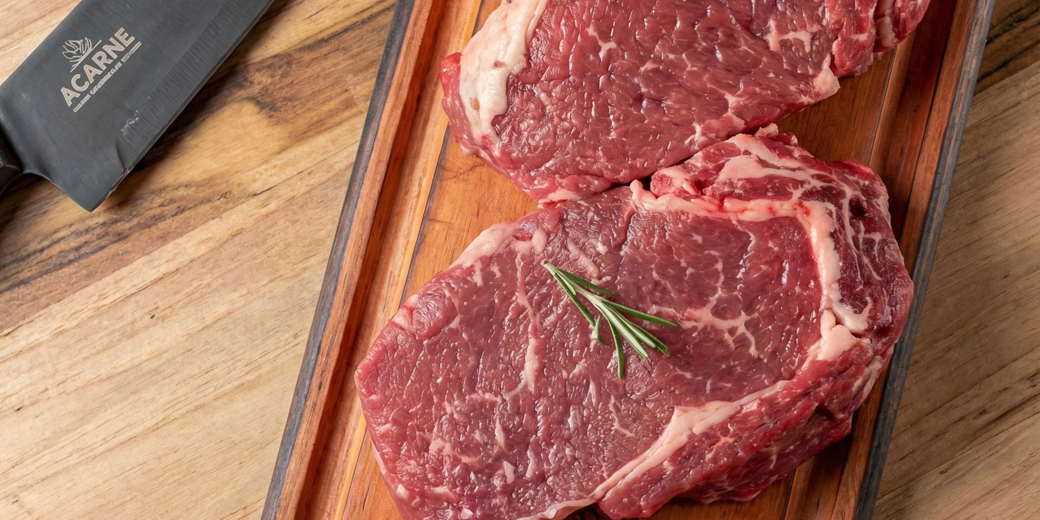 Duas peças altas de bife ancho cruas sobre uma tábua de madeira, exibindo o marmoreio intenso e a gordura entremeada, decoradas com ramos de alecrim ao lado de uma faca