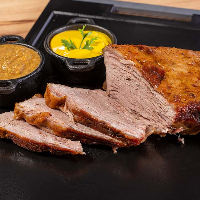 Maminha na Manteiga Fatiada e Pronta Peça de maminha na manteiga assada e fatiada sobre tábua preta, acompanhada de dois molhos em potes pequenos.