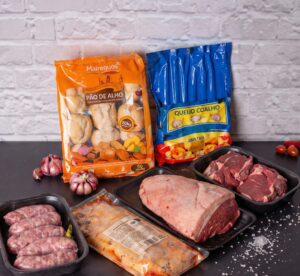 Kit churrasco completo com picanha, linguiça, cortes de bife ancho, pão de alho e queijo coalho dispostos sobre uma mesa preta.