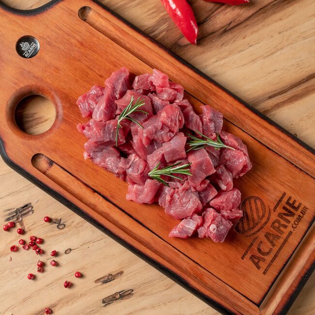 Fotografia overhead de cubos de carne bovina premium frescos e prontos para carne para strogonoff, apresentados em uma tábua de madeira rústica com ramos de alecrim e pimentas-rosa. A tábua exibe a marca ACARNE.com.br, refletindo a qualidade e o frescor que você encontra na Ramalho Carnes para o seu estrogonofe em Campinas.