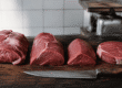 Quatro peças de carne para bife barata alinhadas em tábua de madeira, com faca de açougueiro e balança ao fundo