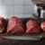 Quatro peças de carne para bife barata alinhadas em tábua de madeira, com faca de açougueiro e balança ao fundo