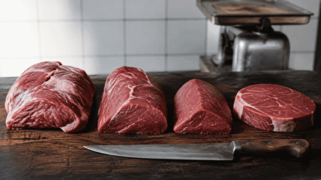 Carne para bife barata: cortes bovinos no balcão do açougue Quatro peças de carne para bife barata alinhadas em tábua de madeira, com faca de açougueiro e balança ao fundo