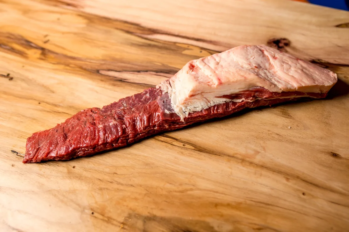 Peça de fraldinha para churrasco crua sobre tábua de madeira, com a fibra longa aparente e a capa de gordura na ponta mais grossa